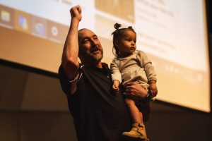 Mr. Steve Lensegrav, holding his granddaughter, is honored on stage in the presence of his family and Principal Tim Wormald.